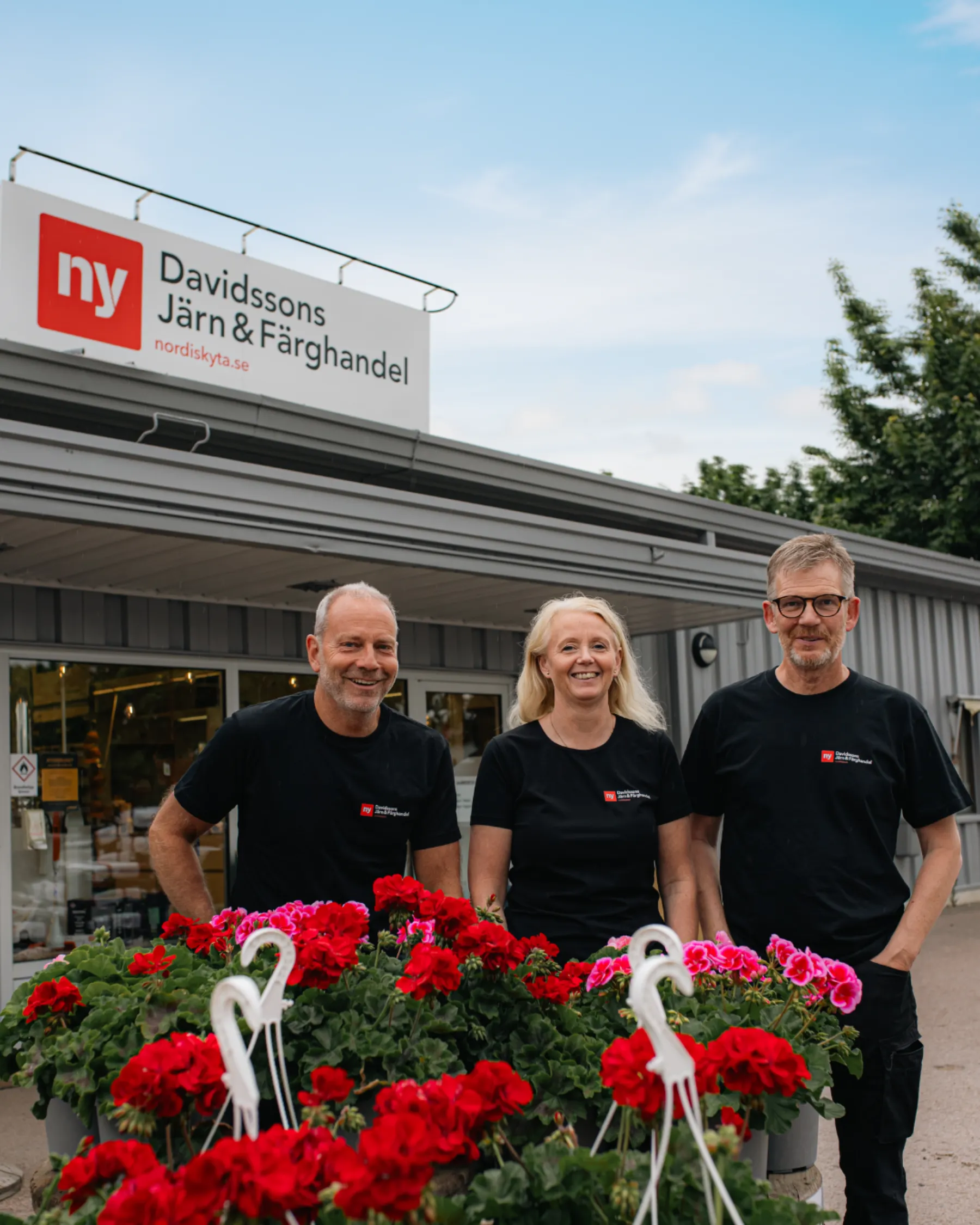 Tre medarbetare från Davidssons Järn & Färghandel framför butiken på Storgatan i Vadstena, med pelargoner i förgrunden.