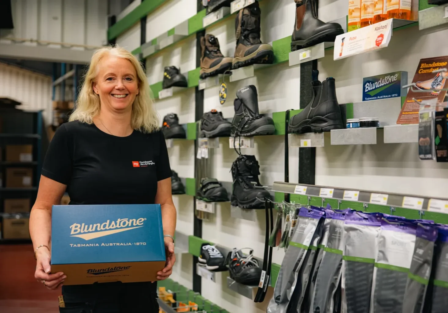 Leende butiksmedarbetare håller en Blundstone skoask framför en vägg med arbetsskor och tillbehör